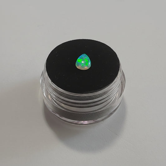 A small teardrop-shaped iridescent gemstone with a green flash displayed on top of a black circular lid of a clear plastic pot against a light grey background.