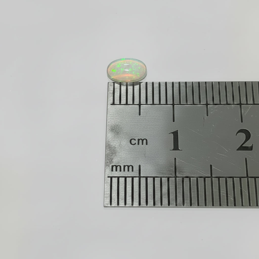 A small clear oval opal In green fire above a metal ruler for scale on a plain light background.