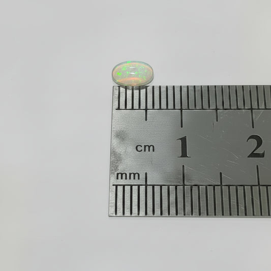 A small clear oval opal In green fire above a metal ruler for scale on a plain light background.