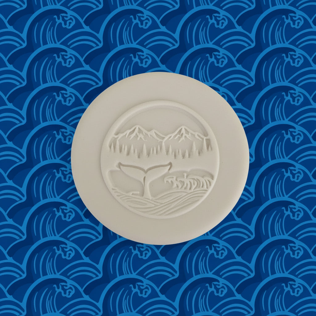 A round clay stamp or with an embossed landscape design featuring mountains, a whale ocean, and waves.