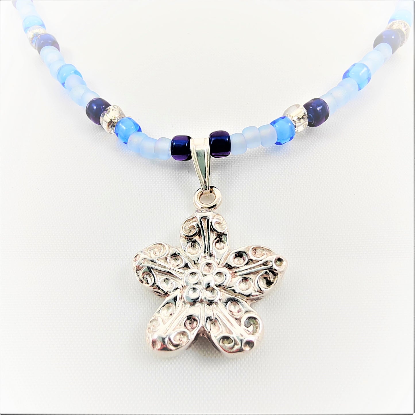 This necklace features a fine silver 999 flower pendant strung on silver glass and blue beads of various hues. Its pendant bail is detachable, allowing for versatility with pendant styles or the addition of other types of pendants. The length of the necklace including flower is approximately 19 inches  The size of flower is 20mm by 18mm  The seed bead sizes are 3mm-4mm