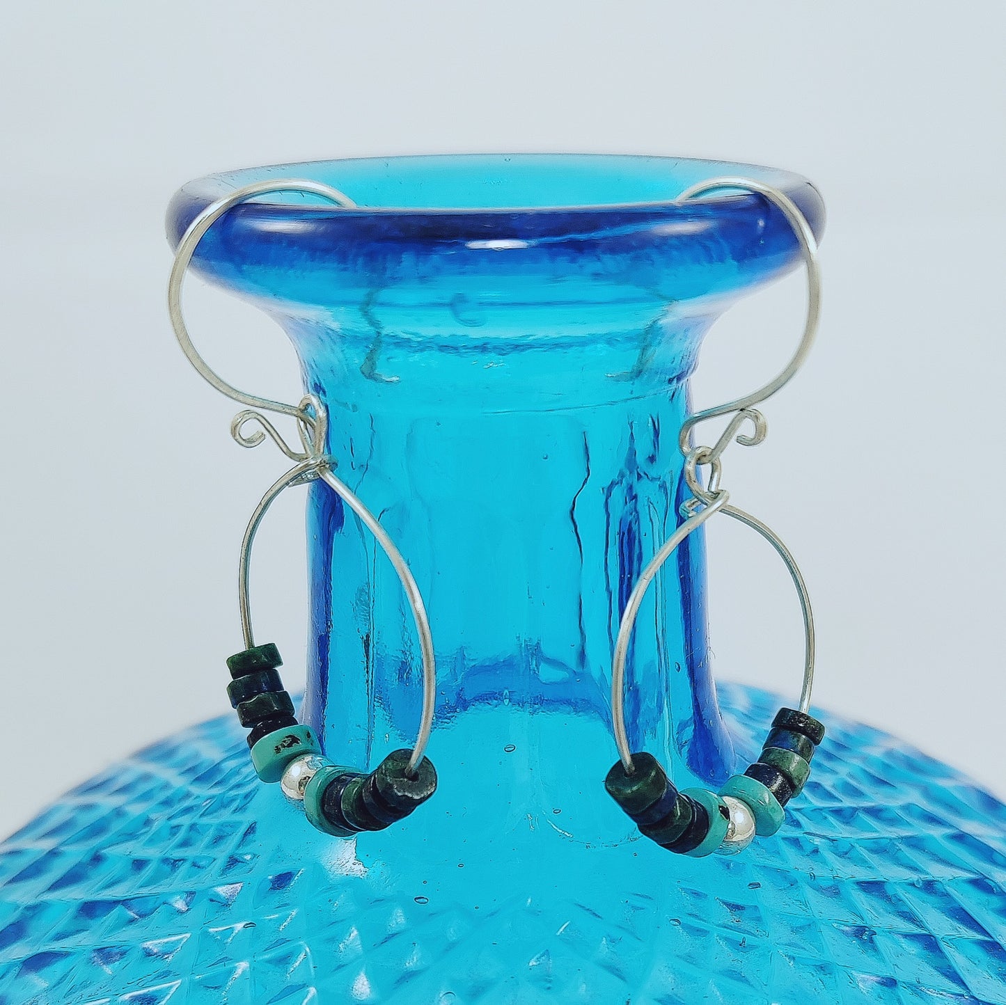 These 925 Sterling Silver hoop earrings with French hook wire feature a scroll detail. The central beads are crafted from heishi beads in shades of green, dark blue, and turquoise, along with a 925 Sterling silver ball. Light and comfortable to wear at only 2.25g each.    Length of earrings are approximately 50mm  Hoop size is 30 x 24mm  Silver ball is 5mm round and the heishi beads are graduated from 6mm x 2mm to 3mm x 2mm.  