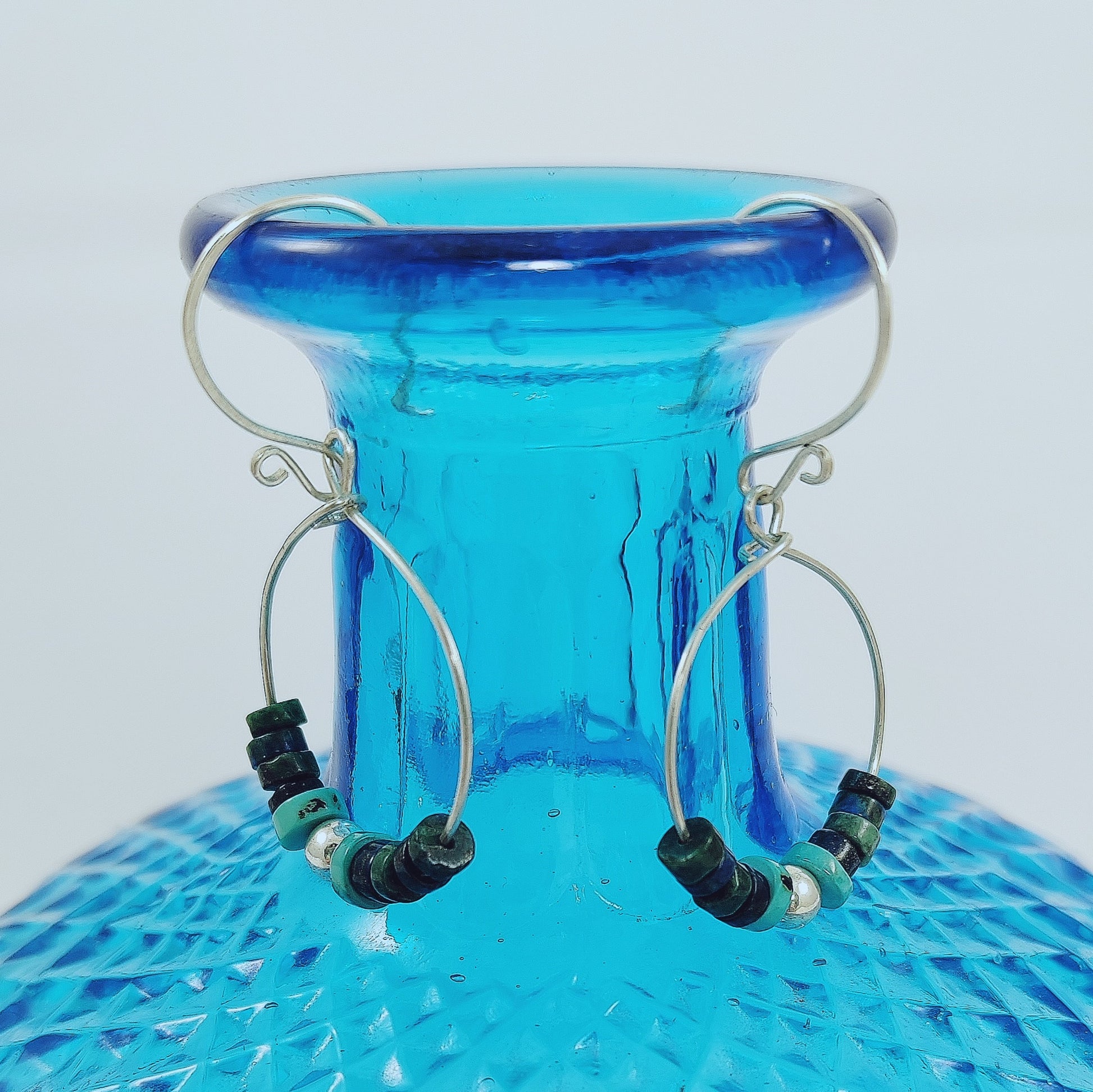 These 925 Sterling Silver hoop earrings with French hook wire feature a scroll detail. The central beads are crafted from heishi beads in shades of green, dark blue, and turquoise, along with a 925 Sterling silver ball. Light and comfortable to wear at only 2.25g each.    Length of earrings are approximately 50mm  Hoop size is 30 x 24mm  Silver ball is 5mm round and the heishi beads are graduated from 6mm x 2mm to 3mm x 2mm.  