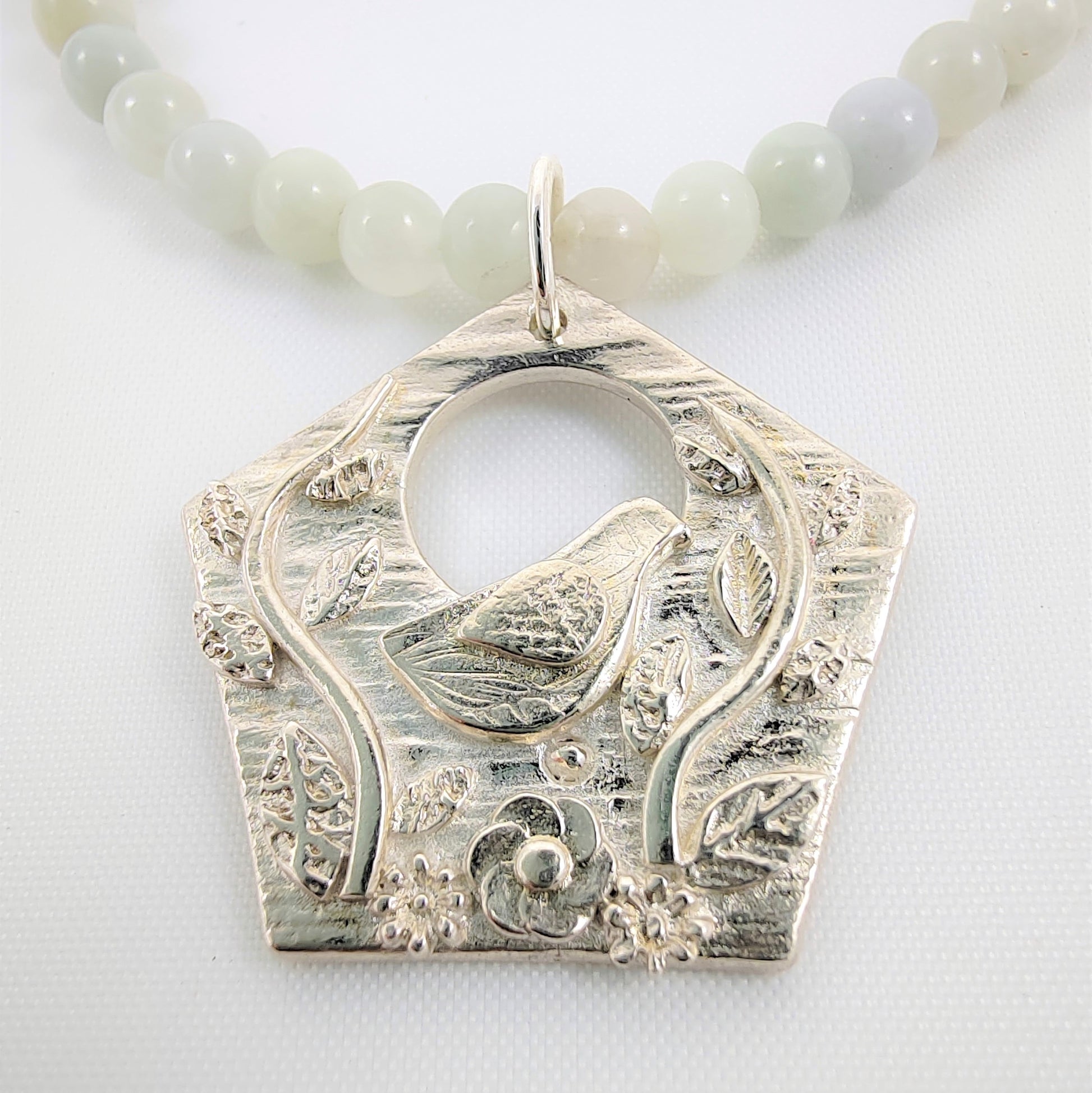 This necklace features a fine silver 999 pendant depicting a bird perched in leafy branches alongside a jade semi-precious beaded necklace attached to a 925 adjustable chain and clasp.