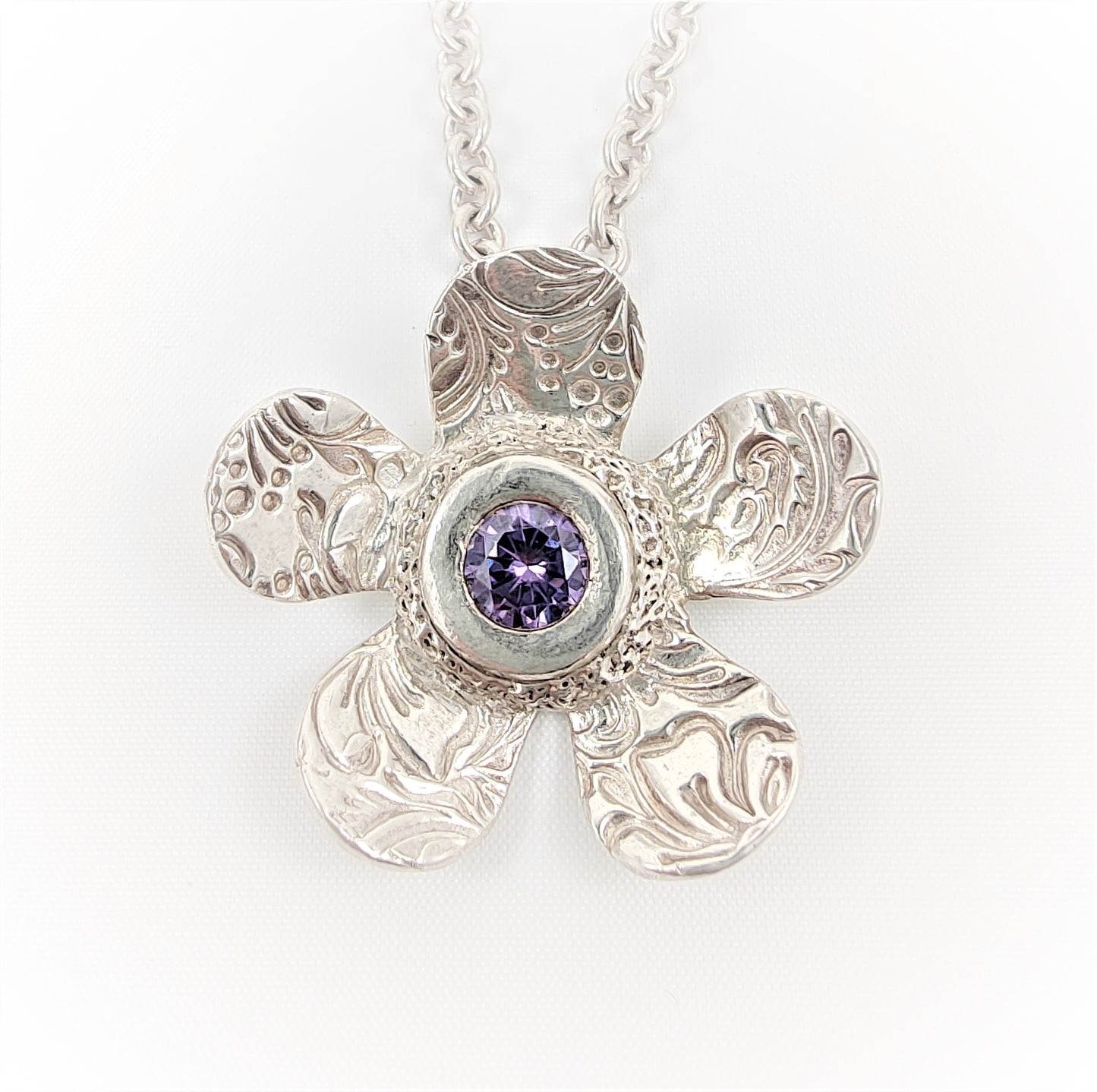 Crafted from heavyweight 999 fine silver, this daisy pendant features a 7mm amethyst-hued cubic zirconia at its centre. It is suspended from a substantial trace chain. The piece is stamped with a full London hallmark on the reverse.