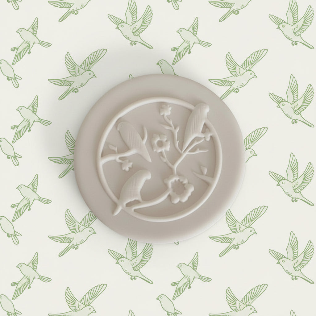 A round clay featuring an embossed design of two birds perched on a flowering branch inside a circle.