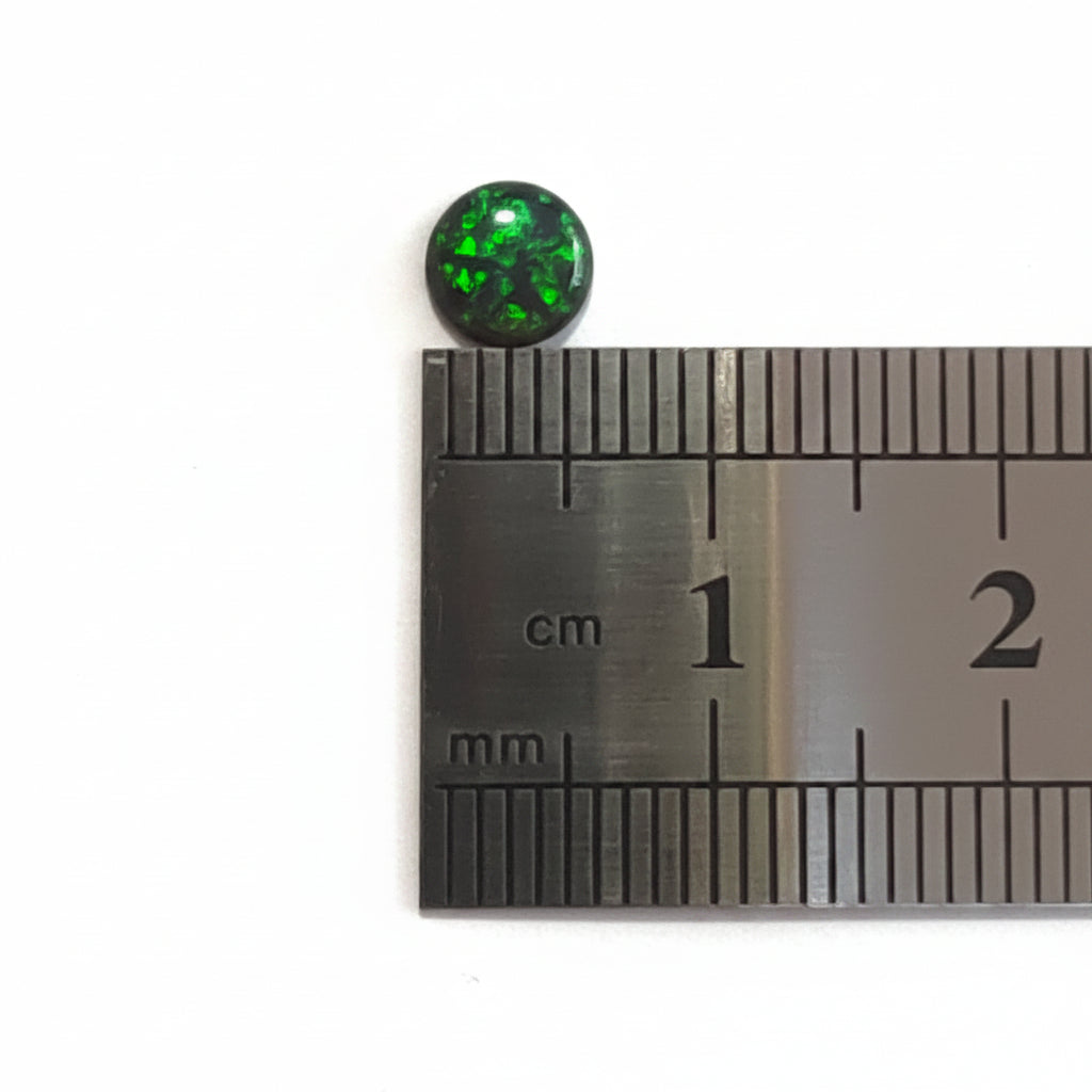 A small round black gemstone with green inclusions, shown next to a metal ruler for scale on a light gray background.