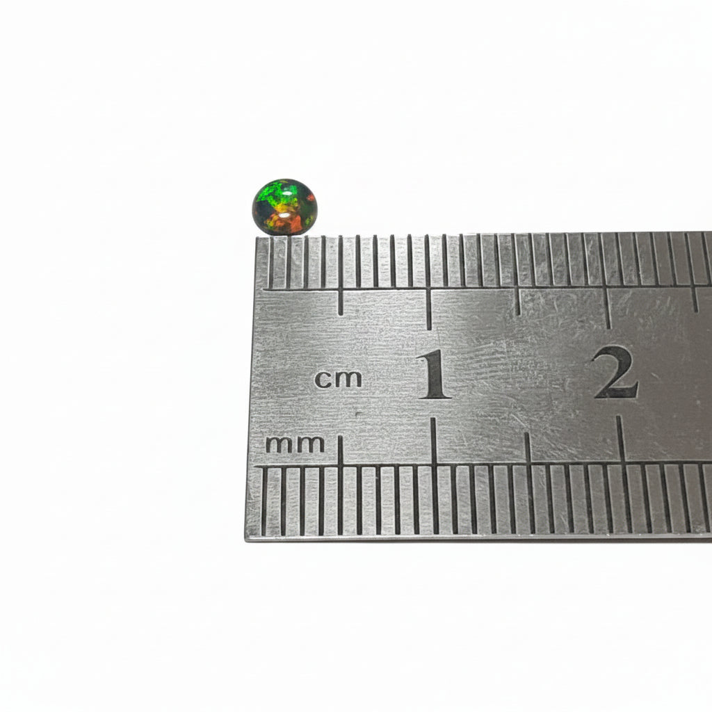 A small round iridescent cabochon resting on a metal ruler for scale, photographed against a plain light background.