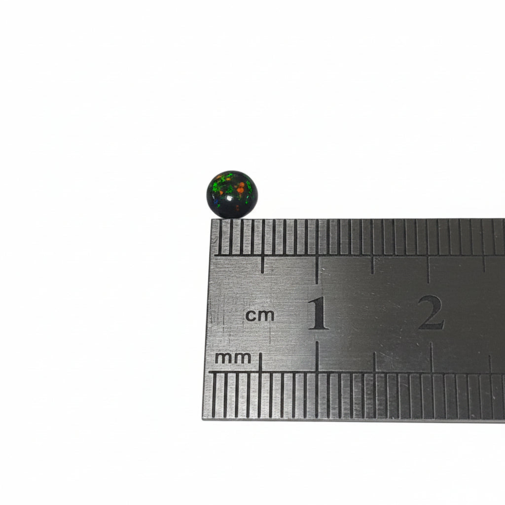 A small round black cabochon gemstone with a multi-coloured opal pattern inside, shown next to a metal ruler for scale on a light background.