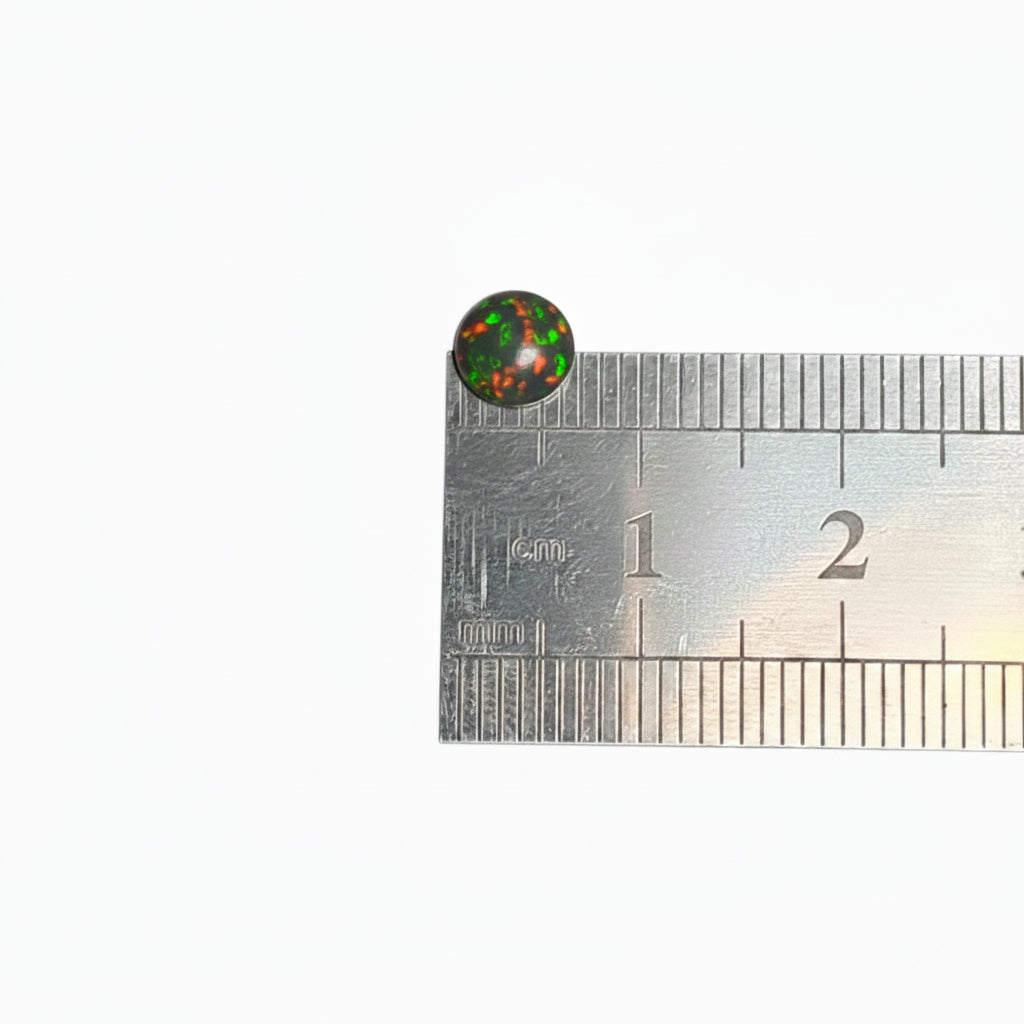A small round black cabochon gemstone with a colorful, iridescent pattern inside, shown next to a metal ruler for scale on a plain light background.