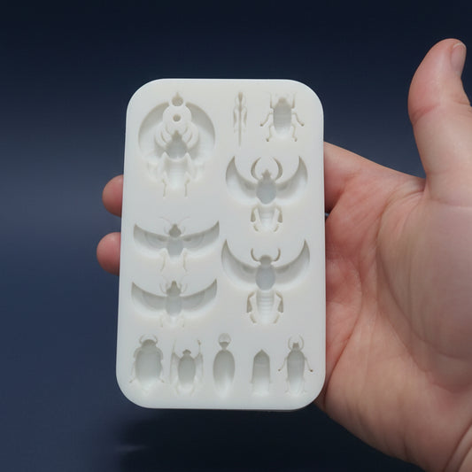 A hand holding a white rectangular silicone mold with multiple insect-shaped cavities, set against a shiny teal background.
