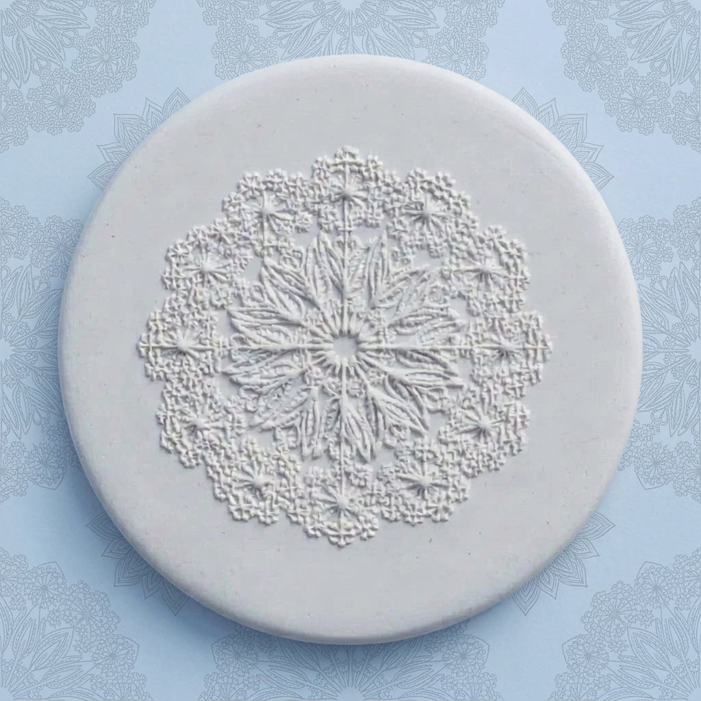 A round, light gray ceramic or stone stamp with an embossed floral mandala design, displayed on a light blue patterned background.
