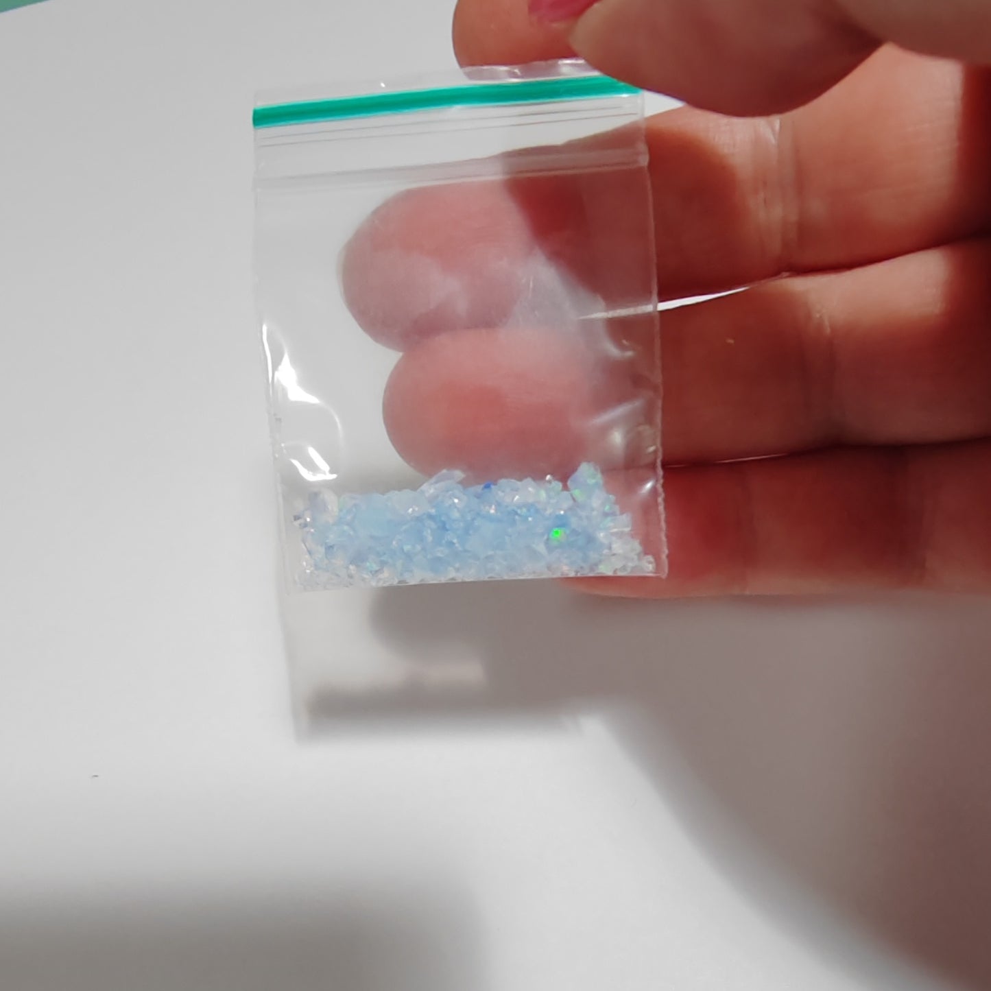 Hand holding a small clear plastic bag with blue fireable crushed opals with green fire on a white background