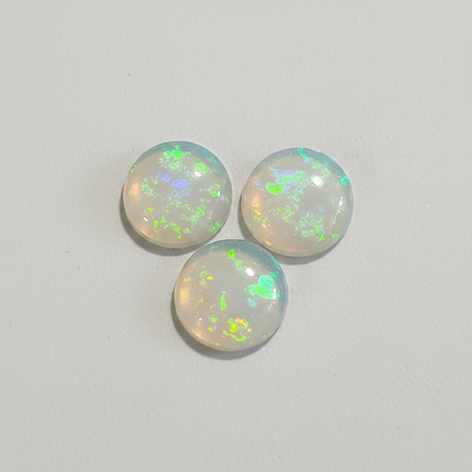 Three round cabochon opal gemstones with a milky white base and vibrant iridescent flashes of green, blue, and orange, arranged in a small triangle on a light gray background.