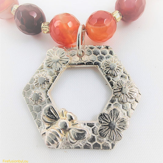 This pendant is crafted from 999 fine silver and 925 findings. It is set with a burnt orange carnelian agate necklace, featuring a Bee Life motif. Showcased is a cheerful bee surrounded by blossoms on a honeycomb texture. The beads are faceted and bi-colored in darker and lighter oranges, with swirls of white, gold foiled seed beads, and spacer beads.