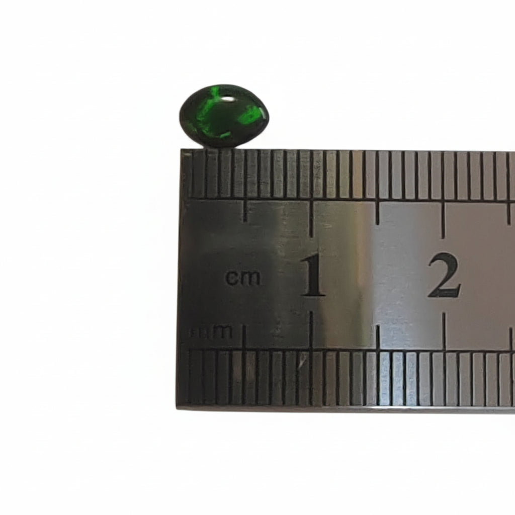 A small marquise green cabochon gemstone placed above a metal ruler for size reference on a plain light background.