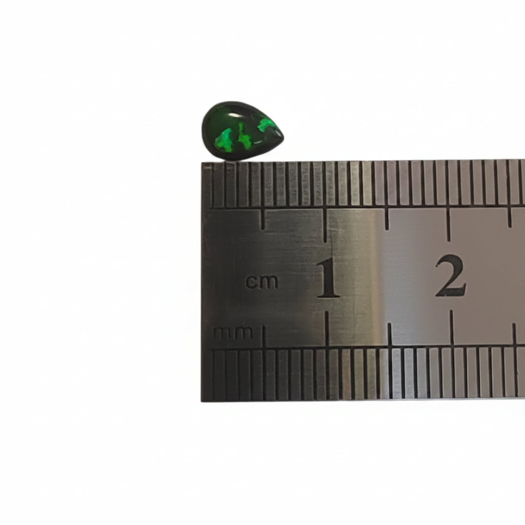 A small teardrop-shaped dark green gemstone with bright green inclusions, shown next to a metal ruler for scale on a light gray background.
