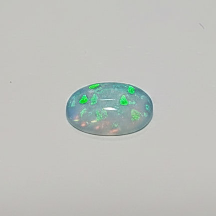A small oval cabochon gemstone with a play-of-colour effect, featuring bright green flashes and blue and white shifts on a neutral grey background.