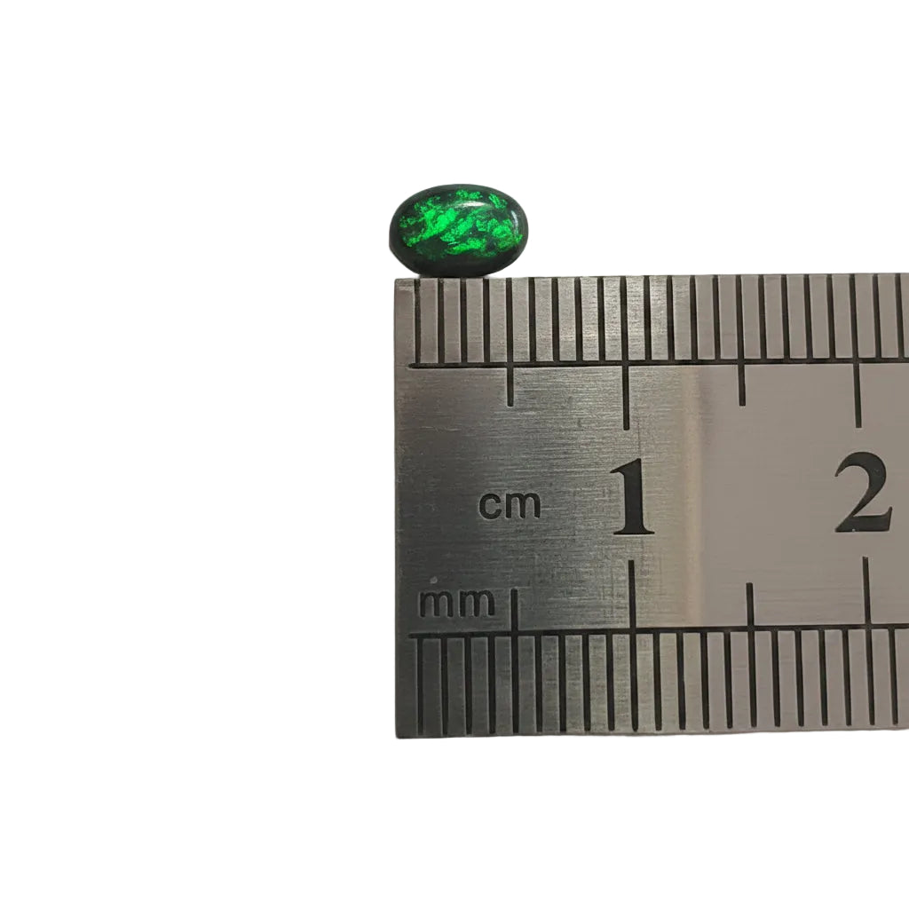 A small oval cabochon gemstone with vibrant green iridescent play-of-color on a black base, shown next to a metal ruler for scale on a plain light background.
