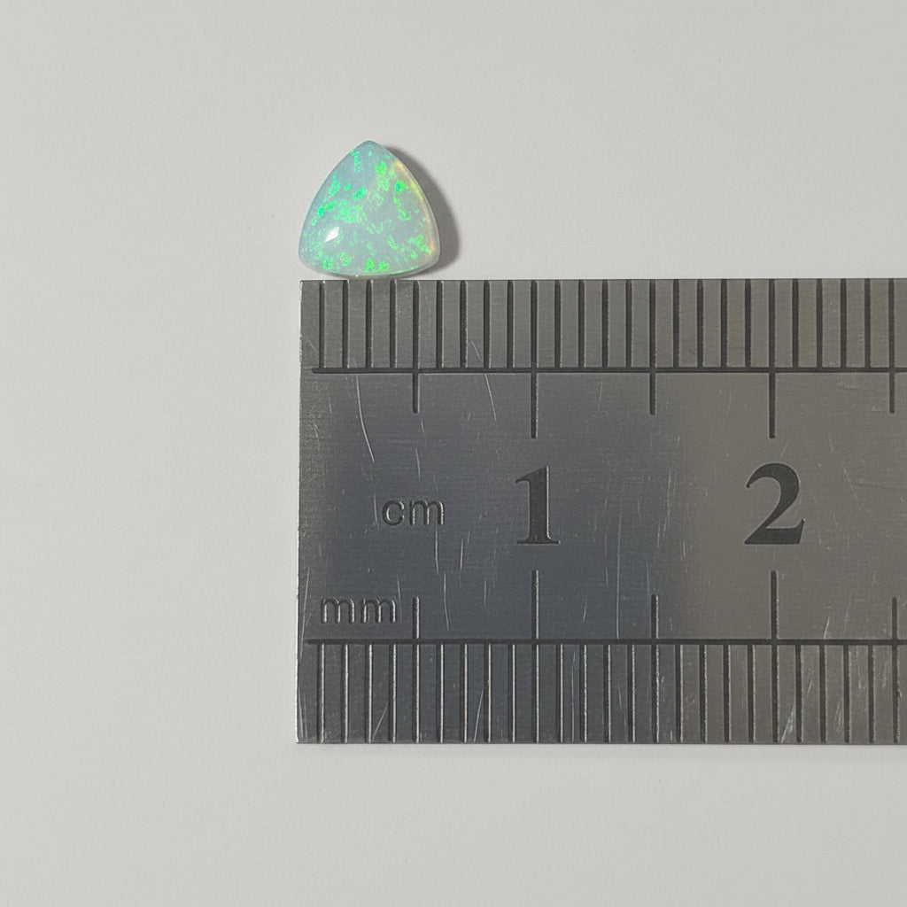 A small trillion-shaped iridescent opal gemstone shown above a metal ruler for scale on a plain light background.