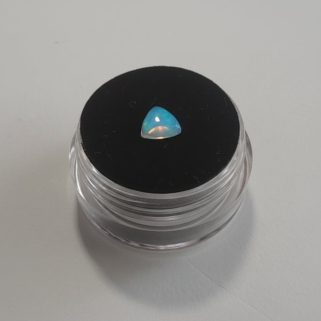 A small triangular iridescent opal gemstone with blue, green, and pink flashes, displayed on a black circular pad inside a clear plastic jar, set against a plain light gray background.
