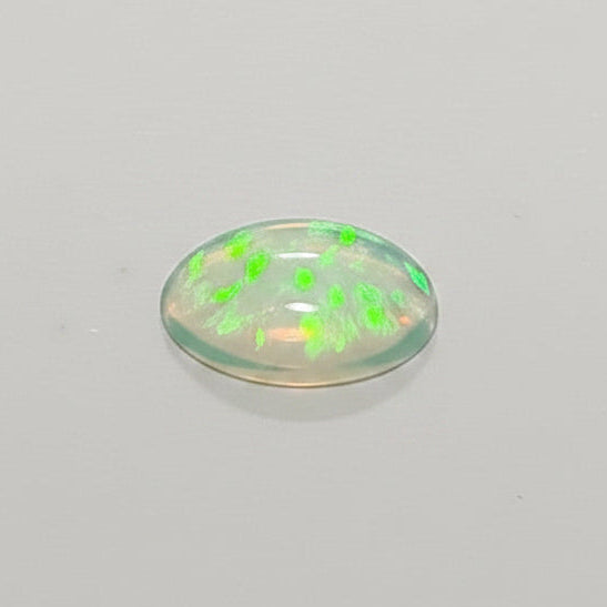A small oval translucent cabochon gemstone with a green flash reflection, shown next to a metal ruler for scale on a plain light background.