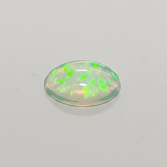 A small oval translucent cabochon gemstone with a green flash reflection, shown next to a metal ruler for scale on a plain light background.