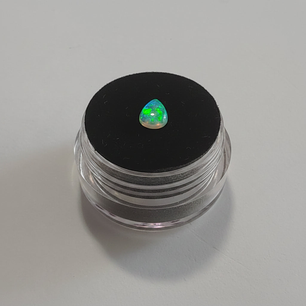 A small teardrop-shaped iridescent gemstone with a green flash displayed on top of a black circular lid of a clear plastic pot against a light grey background.