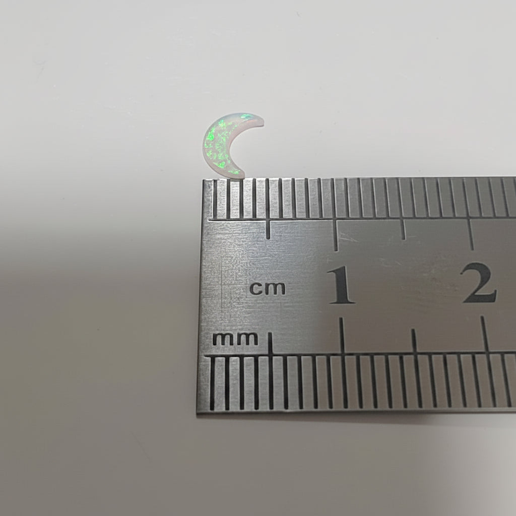 A small iridescent crescent moon-shaped lab grown opal resting on top of a metal ruler with centimetre markings, photographed against a plain light background.