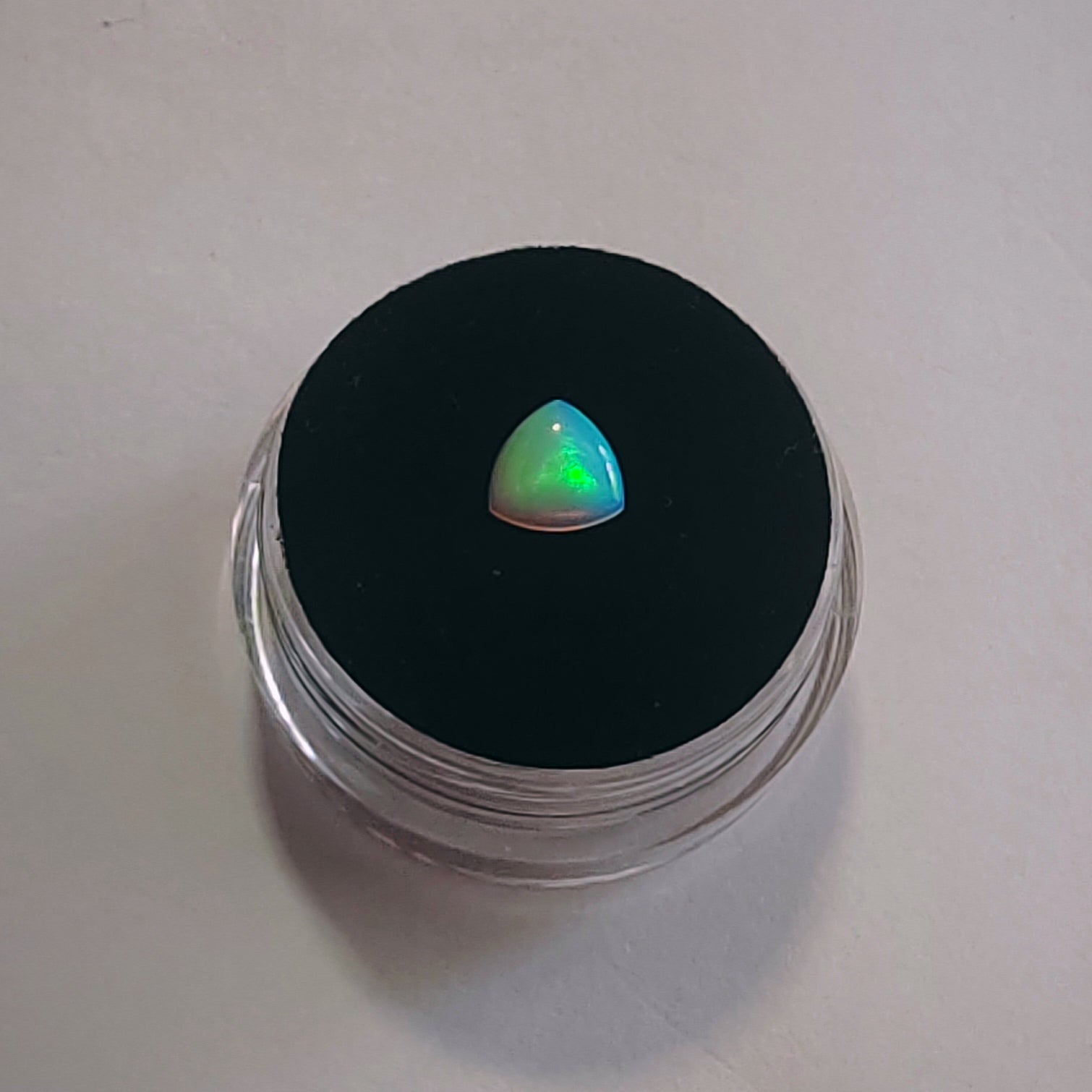 A small triangular iridescent gemstone with green, blue, and pink flashes, displayed on a black circular pad inside a clear plastic jar, set against a plain light gray background.