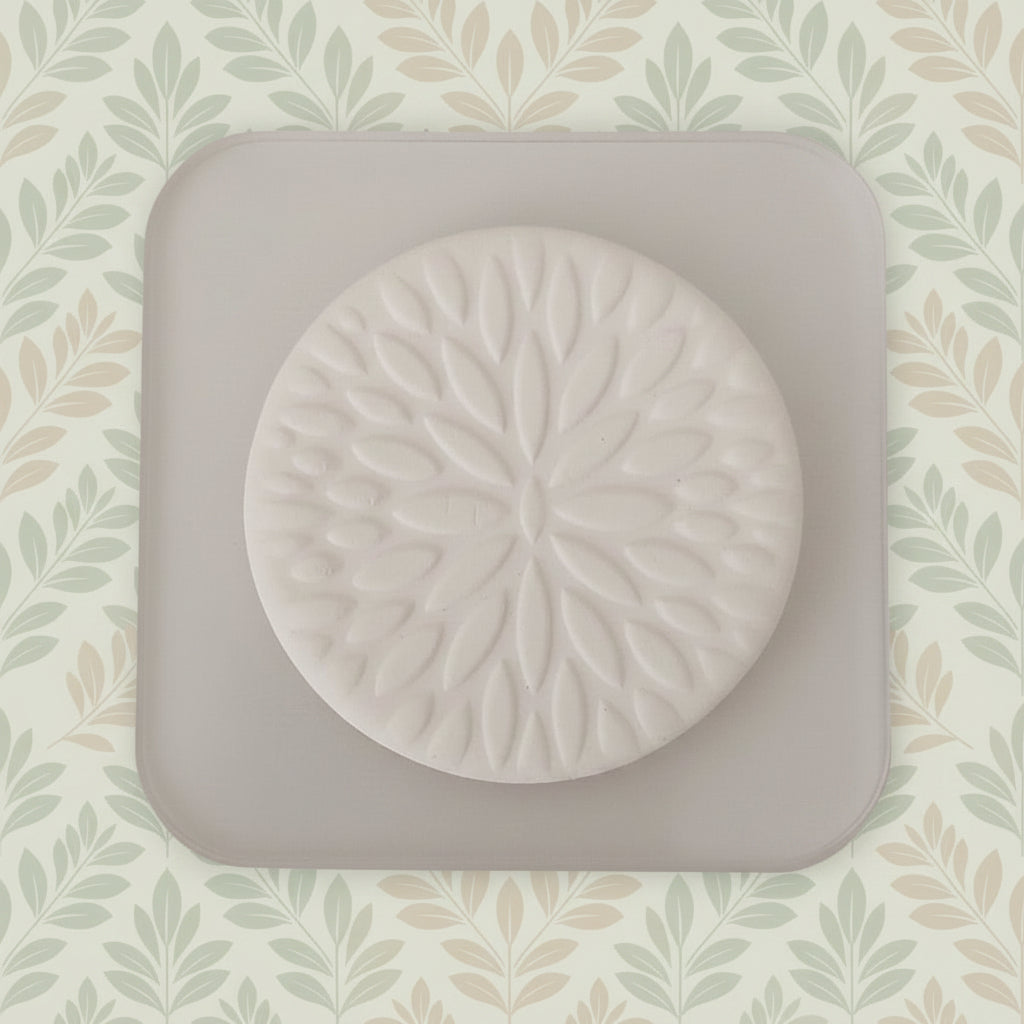 A round clay stamp with a raised floral petal pattern, displayed on a square.