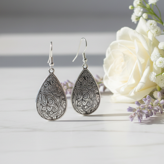 Silver teardrop earrings with intricate patterns on a white surface with white flowers.