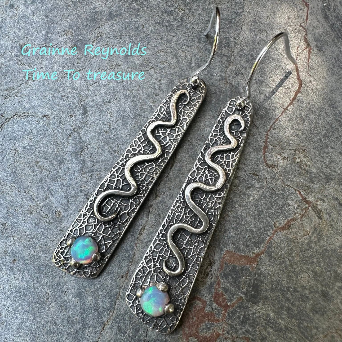 Silver snake-shaped earrings with opal stones on a stone surface, branded 'Grainne Reynolds'.