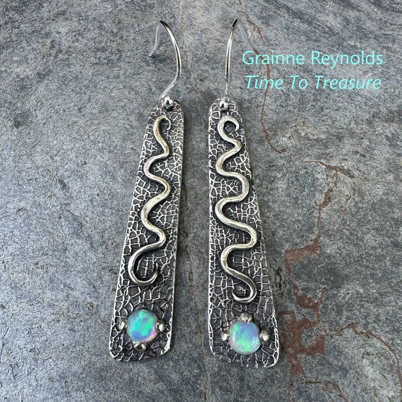 Silver snake earrings with opal stones on a stone surface, branded 'Grainne Reynolds Time To Treasure'.