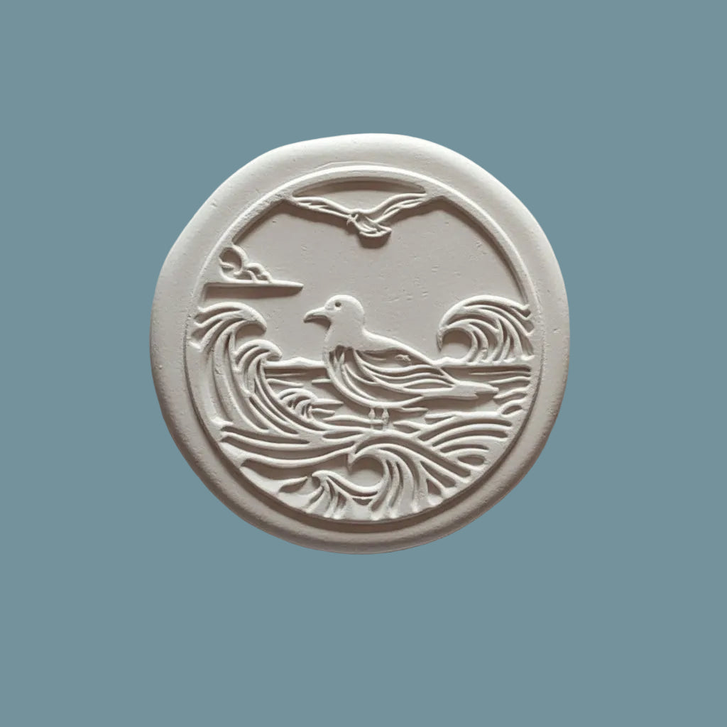 A round  clay stamp featuring an embossed seagull over waves, displayed on a neutral blue-grey surface.