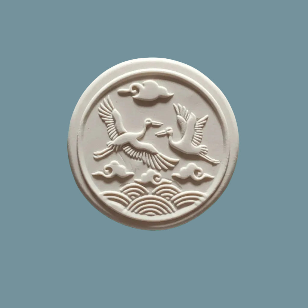 A round  clay stamp featuring embossed cranes flying over waves, displayed on a neutral blue-grey surface.
