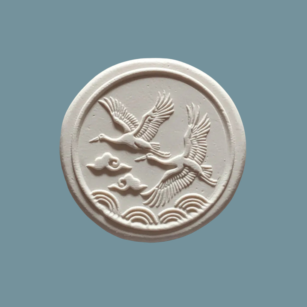 A round  clay stamp featuring embossed cranes flying over waves, displayed on a neutral blue-grey surface.