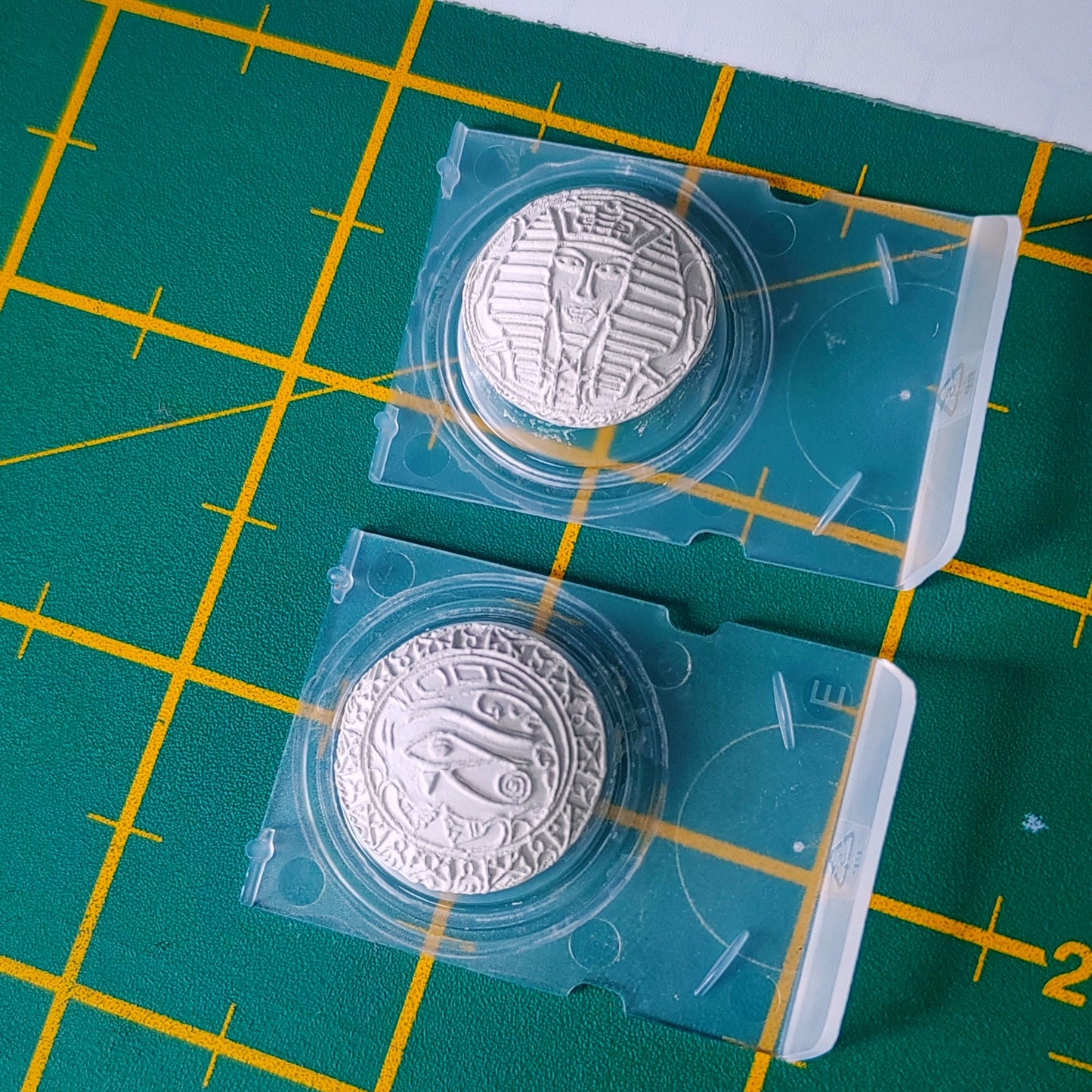 Two clear plastic capsules containing domed silver clay ancient Egypt  with embossed designs, placed on a green cutting mat with yellow grid lines.