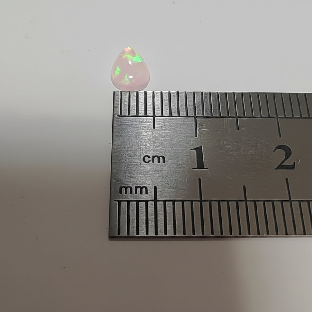 A small teardrop-shaped pink opal gemstone with green and white flashes, shown next to a metal ruler for scale on a plain light background.