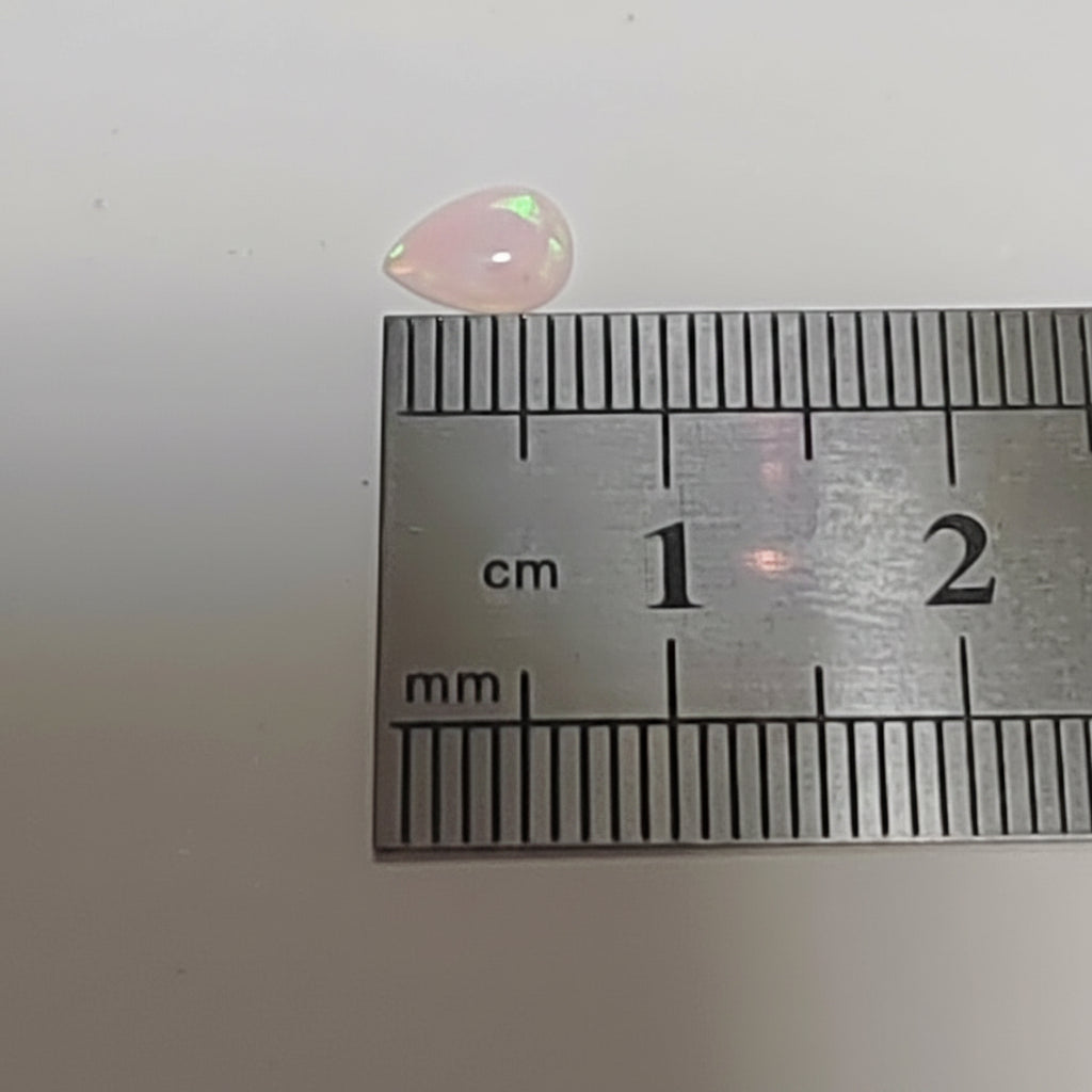 A small iridescent white pink teardrop-shaped gemstone or cabochon placed above a metal ruler for scale, on a plain light background.