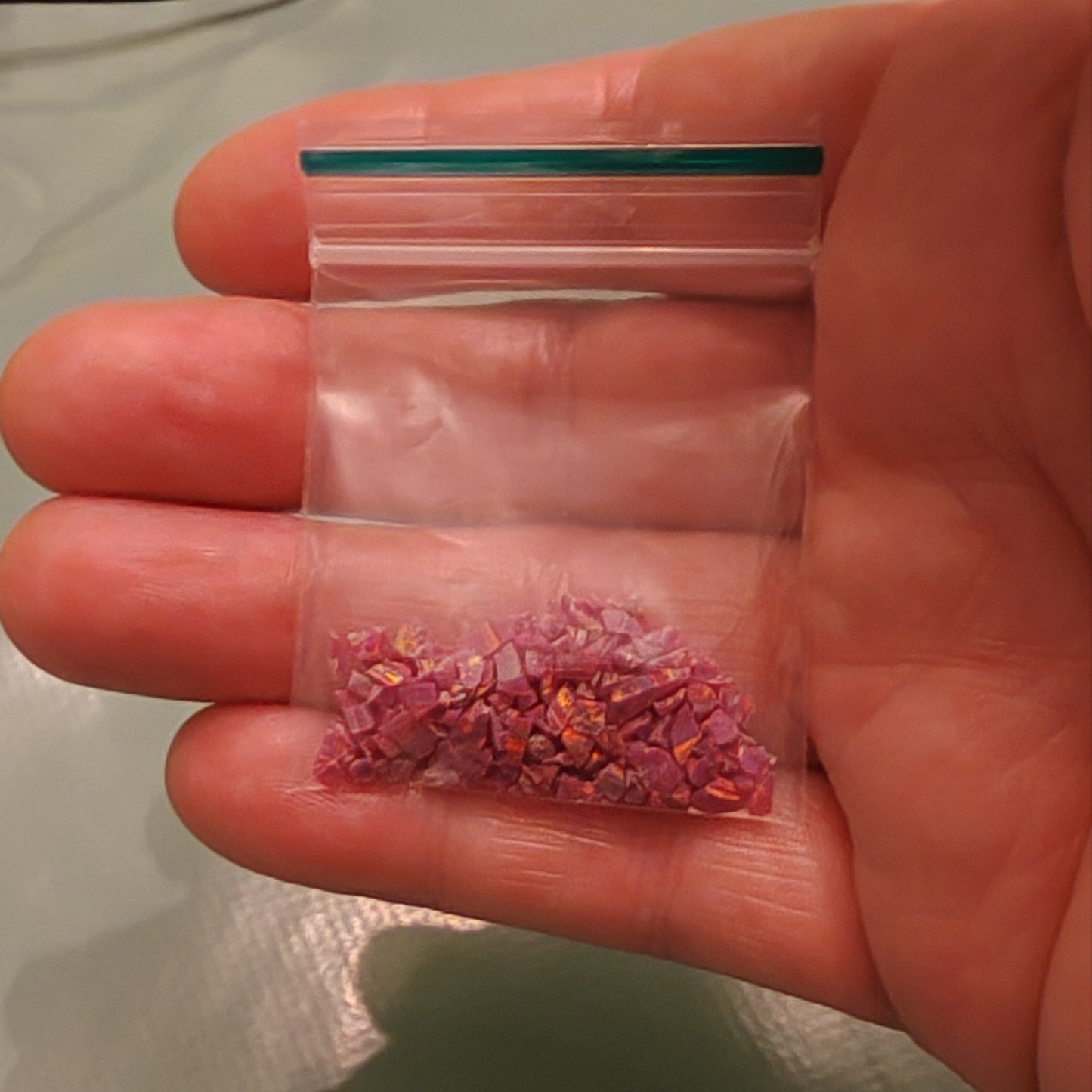 A hand holding a small clear plastic zip-top bag containing crushed pink and purple opal flakes, likely for crafts or jewellery making.