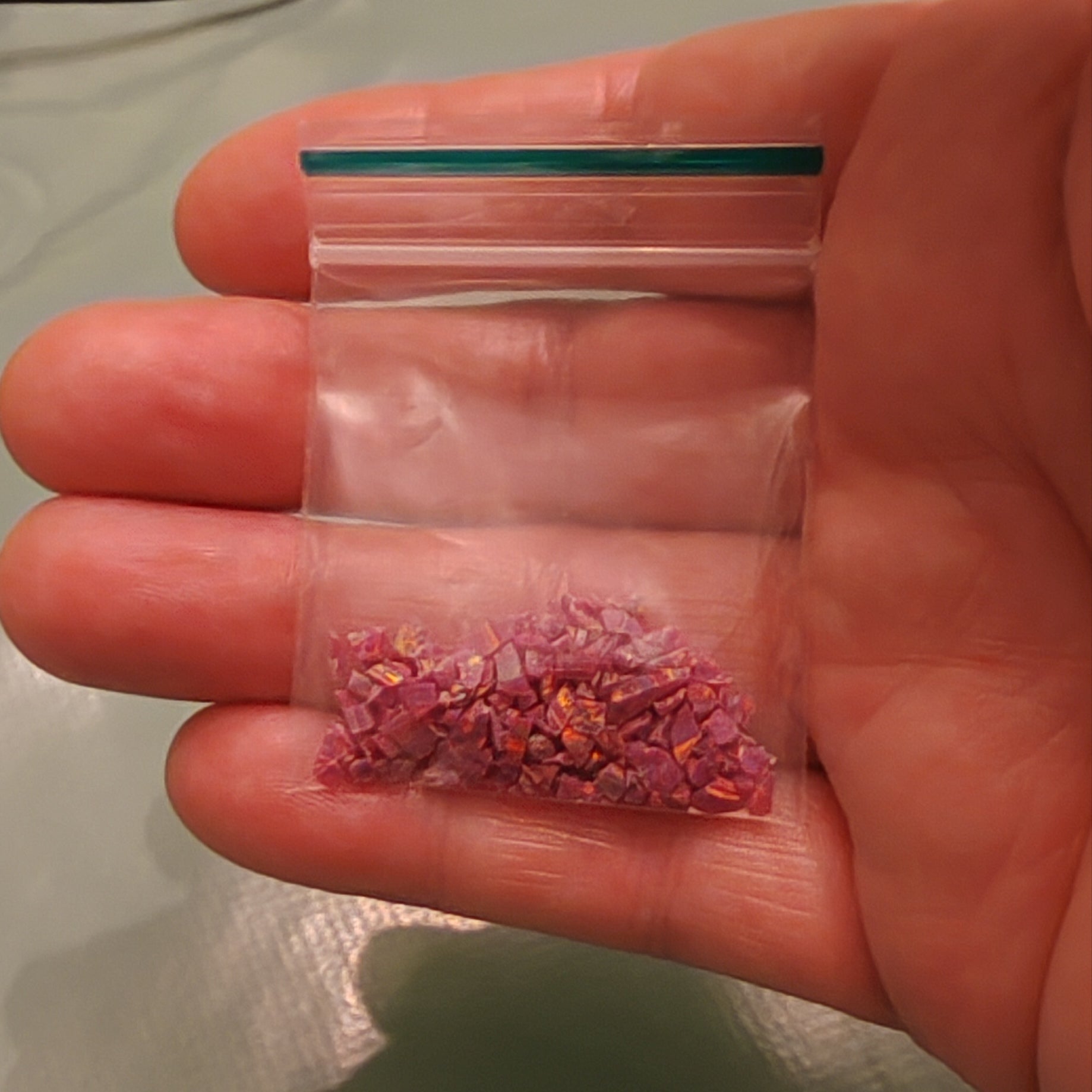 A hand holding a small clear plastic zip-top bag containing crushed pink and purple opal flakes, likely for crafts or jewellery making.