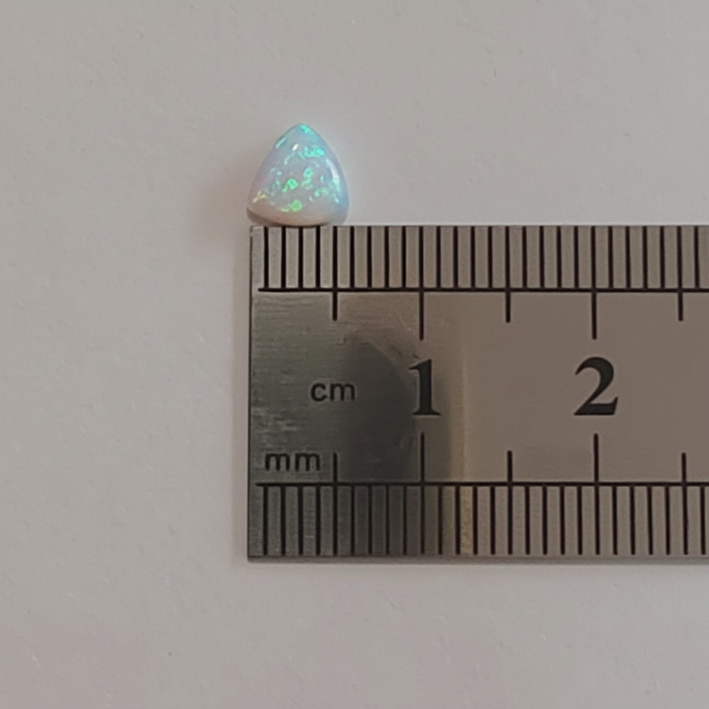 A small iridescent opal cabochon gemstone shown next to a metal ruler for scale on a plain light background.