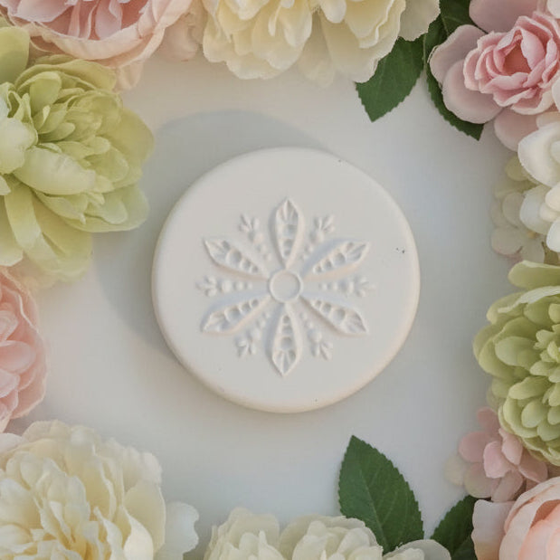 A round clay stamp embossed floral snowflake design, shown on a plain off-white surface.