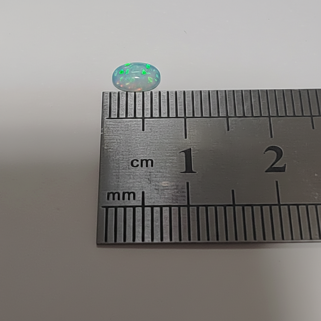 A small oval cabochon gemstone with iridescent green and blue play-of-color, shown next to a metal ruler for scale on a plain light background.