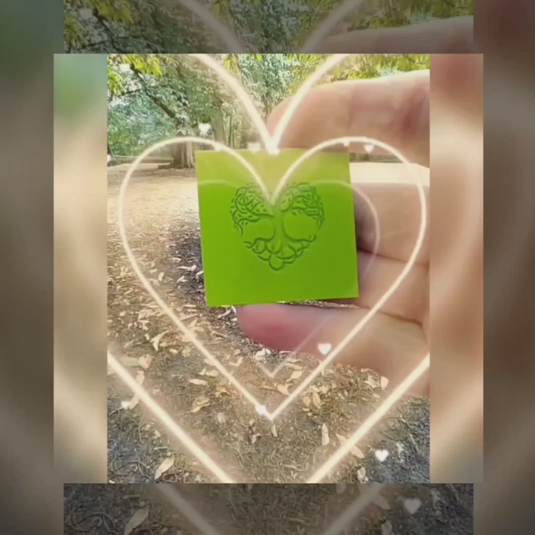 A hand holding a green rubber debossed tree shaped heart with leaves In a Celtic design style, with trees in the background.