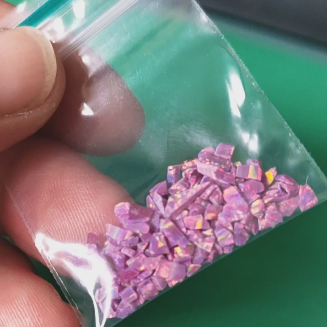 A one gram bag of resin impregnated lab grown opal chips. Raspberry purple colour with gold, pink and green flashes  displayed In a hand.