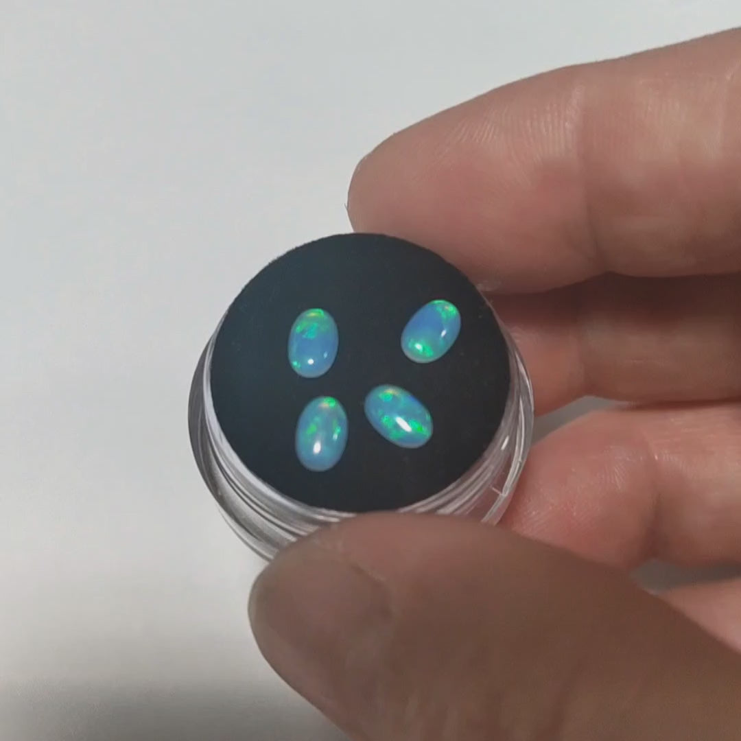 Four blue opals with green fire oval cabochons resting on a black velvet gem box. A hand is showing the green and blue flashes.