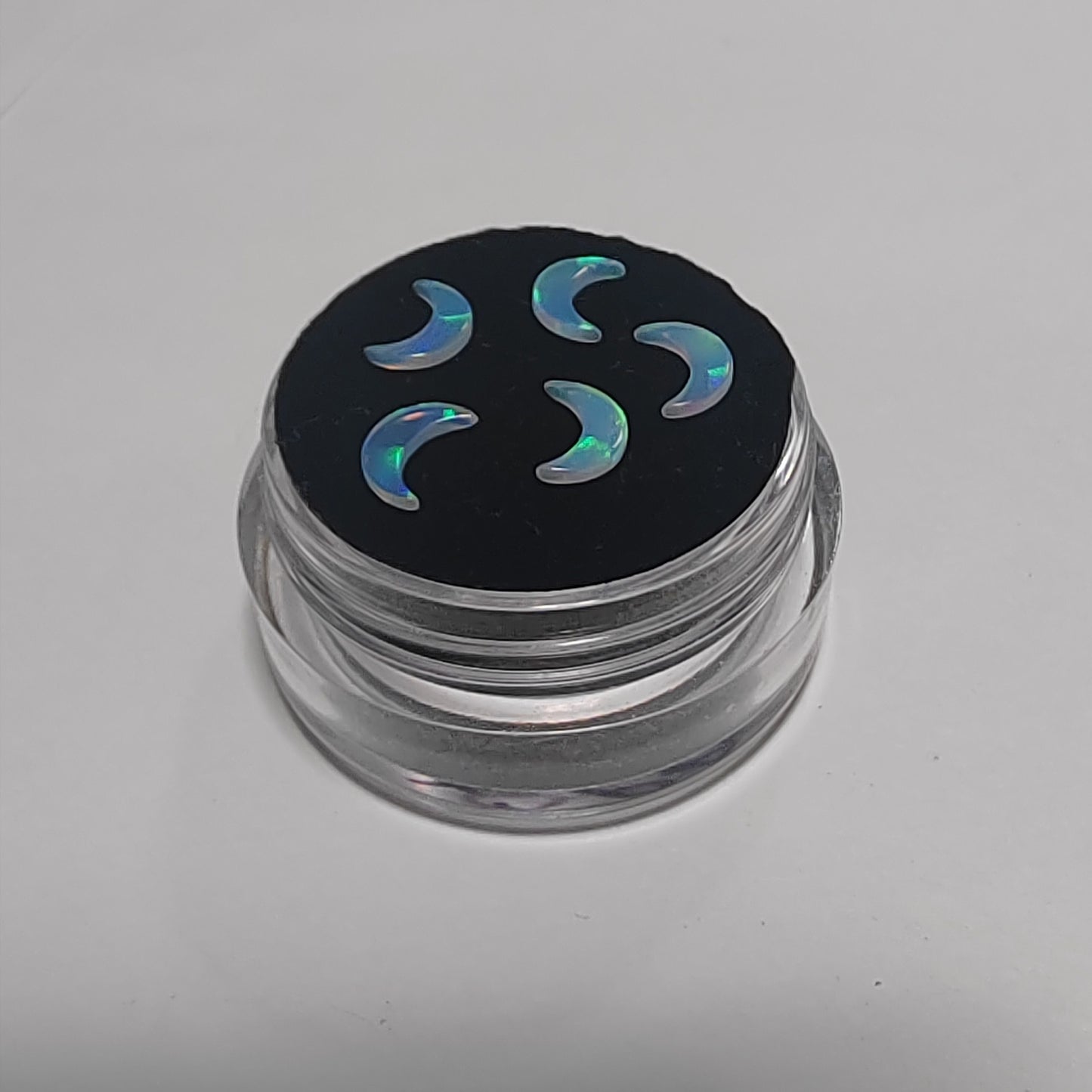 Five crescent-shaped iridescent opal gemstone charms displayed on a black circular insert inside a small clear plastic jar, set against a plain light grey background.