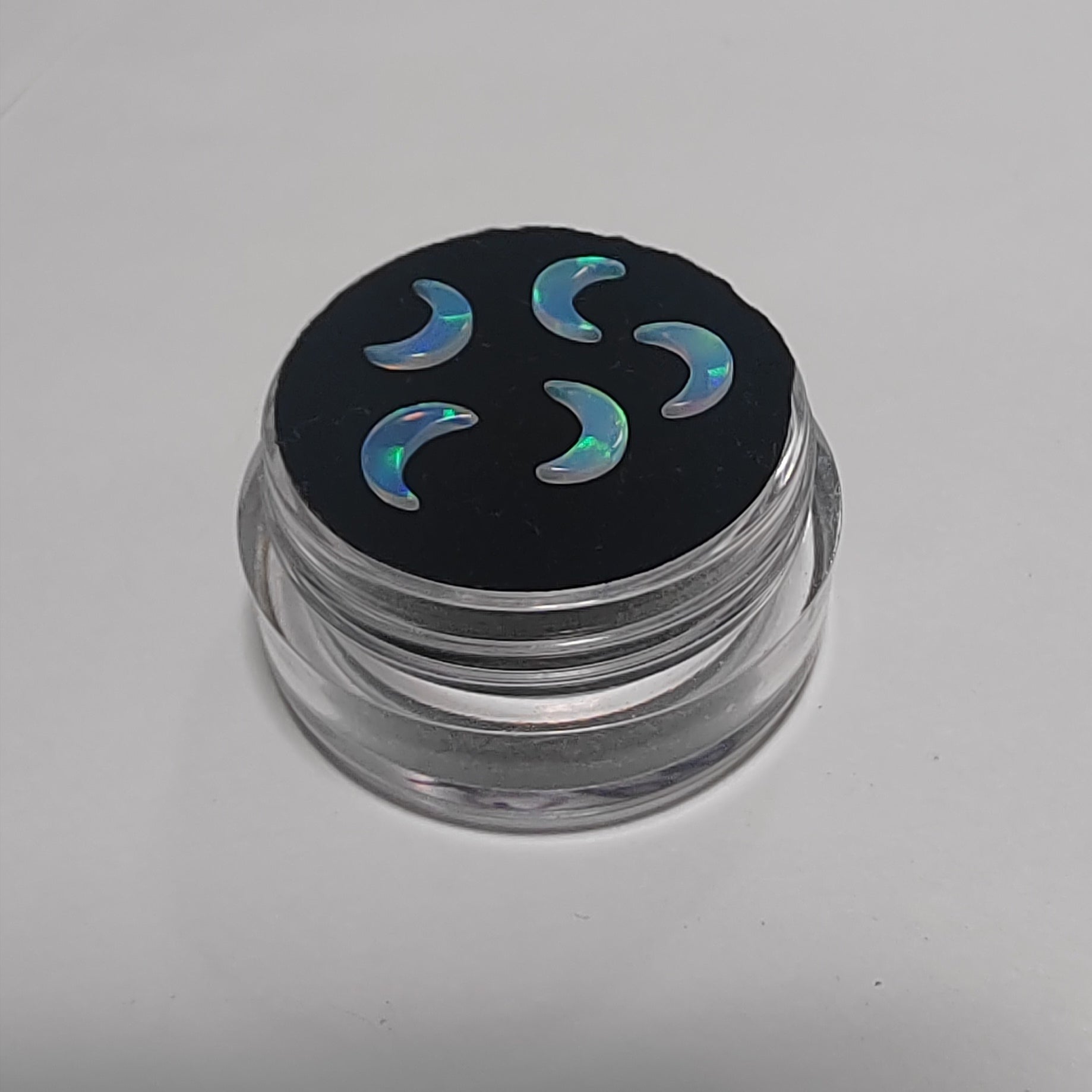 Five crescent-shaped iridescent opal gemstone charms displayed on a black circular insert inside a small clear plastic jar, set against a plain light grey background.
